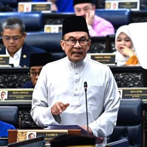 pm anwar announce budget - MKAZ Car Rental - MKAZ Car Rental Malaysian senator speaking at a podium in parliament, wearing a white baju melayu and black songkok, gesturing with his hand.